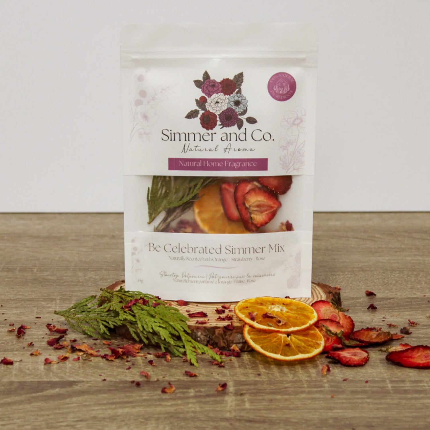pochette blanche contenant un ensemble de pot-pourrie de la marque Simmer and Co. Be Celebrated Simmer Mix avec des tranches d'oranges séchées, des fraises, branches de cèdre sur un fond de texture de bois foncé et l'arrière plan blanc.