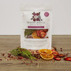 pochette blanche contenant un ensemble de pot-pourrie de la marque Simmer and Co. Be Celebrated Simmer Mix avec des tranches d'oranges séchées, des fraises, branches de cèdre sur un fond de texture de bois foncé et l'arrière plan blanc.