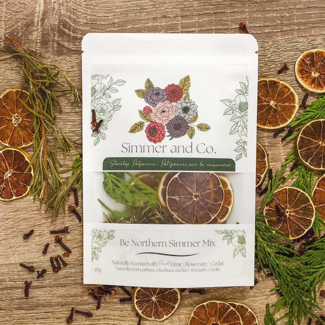 pochette blanche contenant un ensemble de pot-pourrie de la marque Simmer and Co. Be Northern Simmer Mix avec des tranches d'oranges séchées, des branches de romarin et de cèdre à l'intérieur et autour sur un fond de texture de bois brun