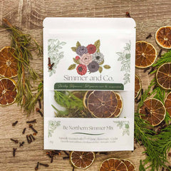 pochette blanche contenant un ensemble de pot-pourrie de la marque Simmer and Co. Be Northern Simmer Mix avec des tranches d'oranges séchées, des branches de romarin et de cèdre à l'intérieur et autour sur un fond de texture de bois brun