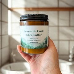 Pot de beurre de karité Eco Loco, 100 % naturel, biologique et équitable, tenu par une main et la personne se trouve dans une salle de bain blanche.