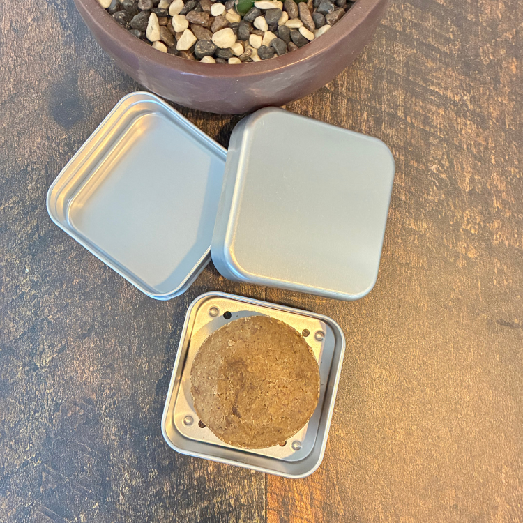 Les 3 étages d'un boitier de métal carré en aluminium avec shampoing solide rond et brun permettant un drainage du shampoing en barre.  Arrière plan d'une planche en bois et d'un pot avec des petites pierres naturelles. 