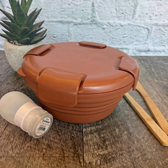 bol en silicone rétractable de couleur rouille avec un couvercle comprenant 4 clips sur le couvercle déposé sur une blanche de bois avec des ustensiles en bois, une petite plante et un arrière plan de brique blanche. 