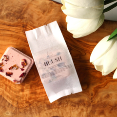 Un sachet blanc écrit blush avec une bombe de bain carré rose sur sa gauche et des tulipes sur  sur sa droite sur une planche de bois. 