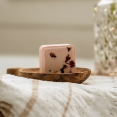 Un bombe de bain carré rose avec des fleurs déposé dans un récipient en bois sur une serviette blanche et beige et avec un fond flou