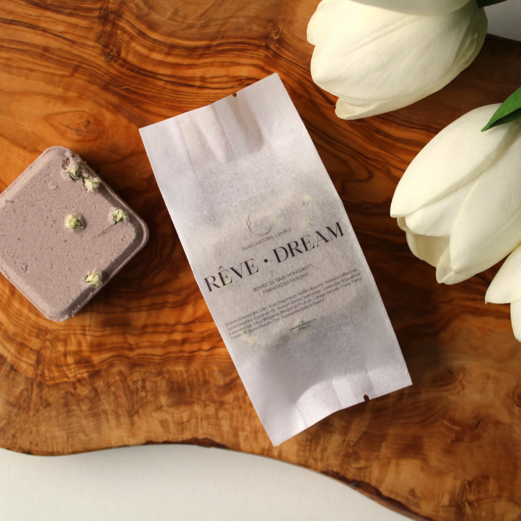 Un sachet blanc écrit Rêve Dream avec une bombe de bain carré vieux rose sur sa gauche et des tulipes sur  sur sa droite sur une planche de bois. 