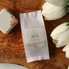 Un sachet blanc écrit zen avec une bombe de bain carré verte sur sa gauche et des tulipes sur  sur sa droite sur une planche de bois. 