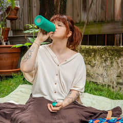 Jeune femme aux cheveux roux qui boit dans une bouteille d'eau réutilisable verte turquoise assise sur une nappe de pique-nique dans un décor de cour de Ville