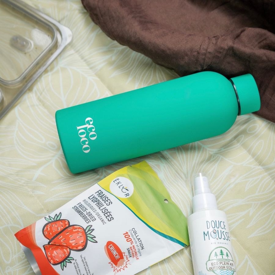 Bouteille d'eau en métal verte turquoise sur une couverture avec un sac de collation fraises lyophilisées et une bouteille de chasse-moustiques Douce Mousse