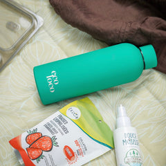 Bouteille d'eau en métal verte turquoise sur une couverture avec un sac de collation fraises lyophilisées et une bouteille de chasse-moustiques Douce Mousse
