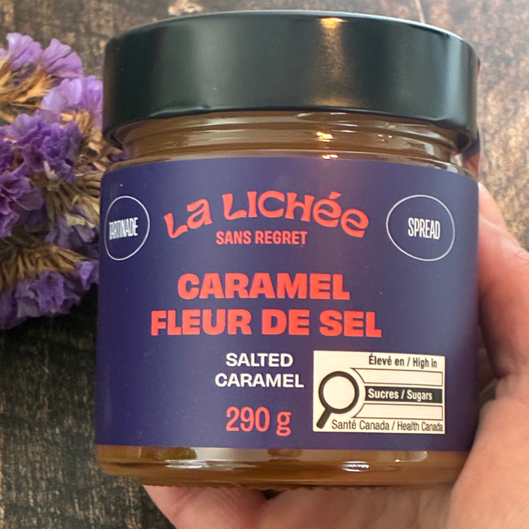 Pot de caramel salé de La liché avec une étiquette mauve tenu dans une main avec une planche de vois et des fleurs mauve en arrière plan. 