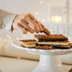 Une main de femme qui prend un morceau de chocolat recouvert de pépites de caramel sur un plateau.  Le chocolat et les pépites de caramel de Caramels FAA sur le plateau sont au chocolat blanc, au chocolat noir et au chocolat au lait. 