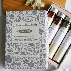 4 fioles au bouchon argenté comprenant 4 lait de bain différents de la marque Doc and Lil devait un coffret cadeau blanc avec des fleurs mauves et une petites fleures blanche sur le dessus déposée sur une planche de bois. 