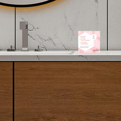 Petite boite rose sur laquelle il est écrit Mélia baume à main déposée sur un comptoir de salle de bain avec une vanité en bois. 