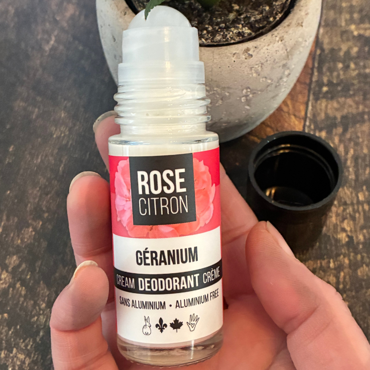 Bouteille de déodorant de la marque Rose  Citron ouverte pour voir la bille, au géranium tenu dans une main et avec une étiquette rouge