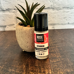Bouteille de déodorant de la marque Rose  Citron au géranium adossé à une petite plante avec une étiquette rouge pour l'odeur de géranium. 