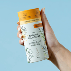 un tube de déodorant blanc et orange tenu dans une main de femme.  On peut y lire déodorant naturel, sans bicarbonate et sans aluminium tanit monoï sur un fond bleu. 