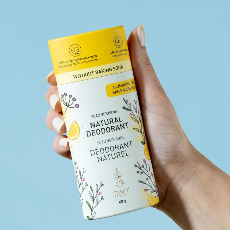 un tube de déodorant blanc et jaune tenu dans une main de femme.  On peut y lire déodorant naturel, sans bicarbonate et sans aluminium tanit yuzu et vervaine sur un fond bleu. 