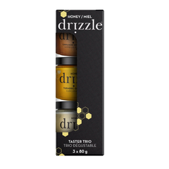 Emballage noir de 3 miels de la marque Drizzle.  Nous voyons qu'une partie des pots dans l'emballage et ceux-ci sont blanc, couleur curcuma et couleur cannelle sur un fond blanc. 