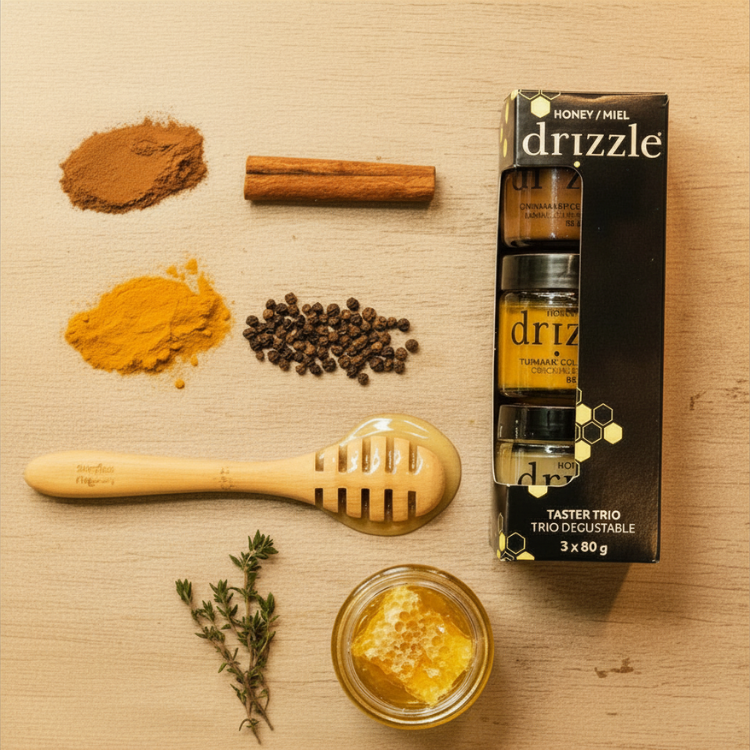 Une ensemble de trois pots de miels de différentes couleurs dans un emballage en carton noir de la marque Drizzle.  À gauche de l'ensemble, on voit de la cannelle, du poinvre, du curmuma, une cuillère à miel et un pot contenant du miel. 