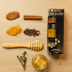 Une ensemble de trois pots de miels de différentes couleurs dans un emballage en carton noir de la marque Drizzle.  À gauche de l'ensemble, on voit de la cannelle, du poinvre, du curmuma, une cuillère à miel et un pot contenant du miel. 
