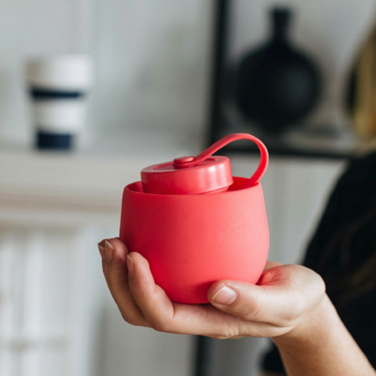 Une gourde rouge en silicone rétractable montrant l'effet lorsque celle-ci atteint son format ne fois rétracté, tenu dans une main de femme avec un fond flou majoritairement blanc. 