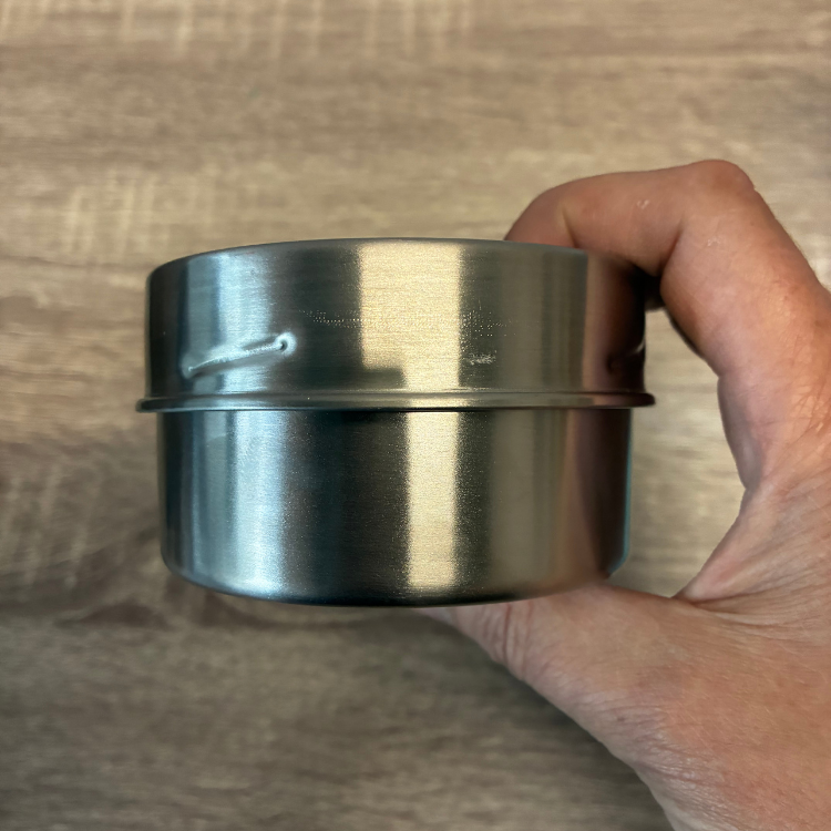 Un  pot avec un couvercle en acier inoxydable argent enu dans une ain de femme au-dessus d'une planche en bois. 