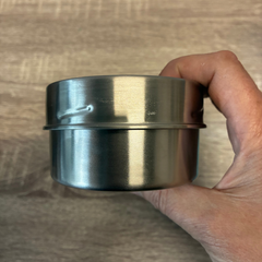 Un  pot avec un couvercle en acier inoxydable argent enu dans une ain de femme au-dessus d'une planche en bois. 