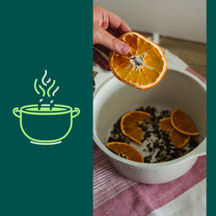 Une main qui met une tranche d'orange séchée dans un chaudron blanc qui contient des oranges séchés, des branches et herbes déposé sur un linge à vaisselle carrelé blanc et rouge. contour de l'image verte forêt avec une illustration de chaudron qui fume vert lime.