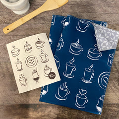 Un linge à vaisselle en microfibre à motifs de tasses sur fond bleu et un chiffon suédois beige à motifs de tasse à café sur un comptoir de bois foncé