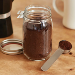 Un pot en verre contenant du café moulu avec une cuillère en acier inoxydable et un couvercle, déposé sur un comptoir en bois. 