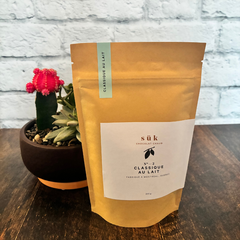 Sac de chocolat chaud classique au lait de SÜK une entreprise québécoise sur une planche de bois avec une plante en arrière plan. 