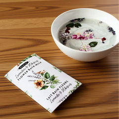 Une sachet rectangulaire blanc écrit lait de bain fleure lavande et hibiscus en francais et en anglais et le logo de Dot & Lil avec un bol d'un liquide laiteux avec de fleur sur un fond de planche en bois. 