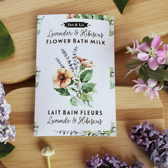 Une sachet rectangulaire blanc écrit lait de bain fleurs Lavande et hibiscus en francais et en anglais et le logo de Dot & Lil sur une planche en bois, avec une des lilas et autre type de fleurs. 