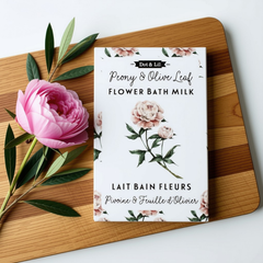 Une sachet rectangulaire blanc écrit lait de bain fleure pivoine et feuille d'olivier en francais et en anglais et le logo de Dot & Lil sur une planche en bois, avec une pivoine rose et des feuilles d'olivier sur fond blanc. 