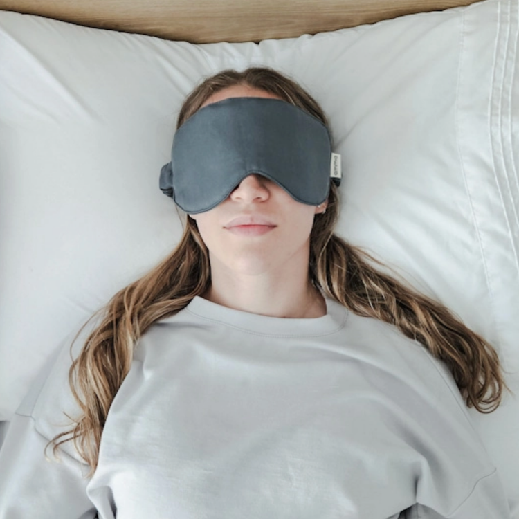 Une femme au cheveux long chatain couché sur le dos la tête apposée sur un oreiller blanc portant un masque de sommeil gris de la marque Amma