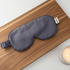 Un masque de nuit gris foncé de l marque Amma sur une planche de bois accompagné d'une chandelle dans un pot de ciment. 