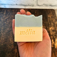 Le savon de la marque Mélia des Miels d'Anicet de couleur bleu et crème au géranium et à la menthe tenu dans une main au dessus d'une planche de bois. 