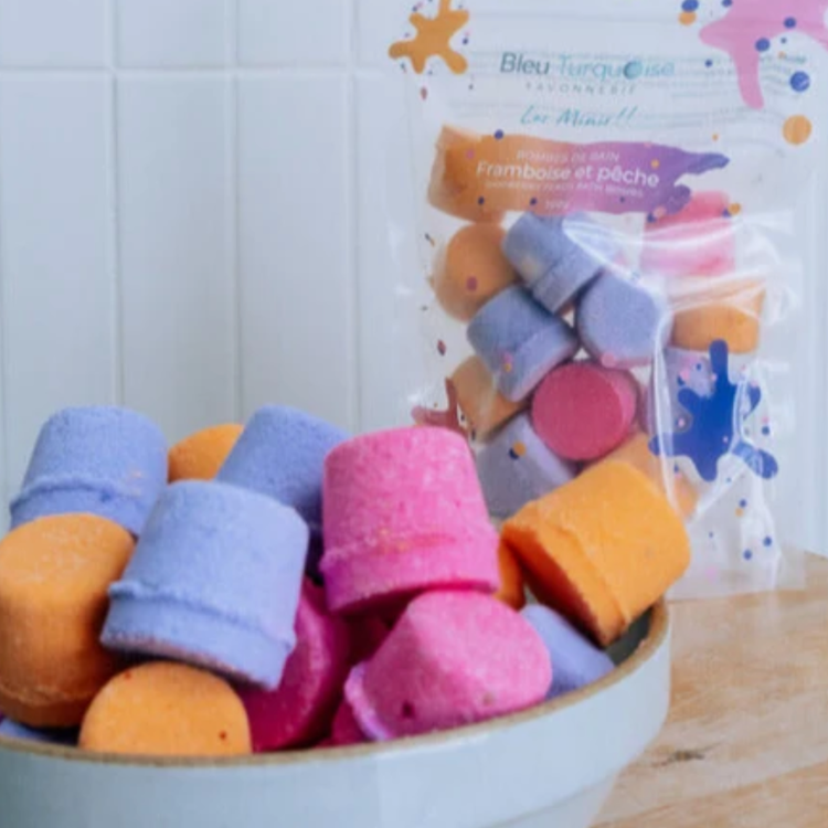Mini bombes de bain en forme de bouchon multicolores bleu, rose et orange dans un pot blanc avec un sac de Bleu Turquoise savonnerie en arrière plan. 