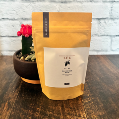 Sac de chocolat chaud classique noir de l'entreprise québécoise SÜK sur une planche de bois avec une plante en arrière plan. 