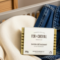 Un savon vert avec des stries et une étiquettes ou l'on peut lire:  Fer a cheval, Marseille, Savon détachant, stain remover, savon de marseille 300 déposé dans un panier de vêtement dans lequel il y a un tricot blanc et un jean. 