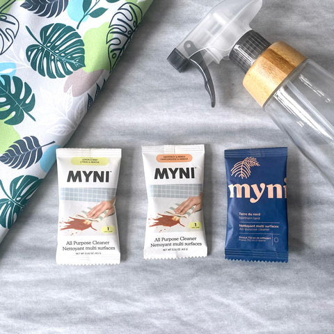 Trois sachets bleus pâle et blanc de la marque Myni contenant une pastille de nettoyant multi usage sur un fond de textile gris et un linge pour nettoyer les vitres à motifs de feuilles tropicales ainsi qu'un pulvérisateur gris noir et manchon de bois.