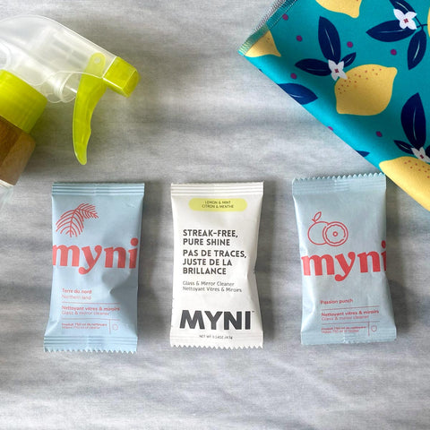 Trois sachets bleus pâle et blanc de la marque Myni contenant une pastille de nettoyant à vitre sur un fond de textile gris et un linge pour nettoyer les vitres à motifs de citrons ainsi qu'un pulvérisateur vert et manchon de bois.