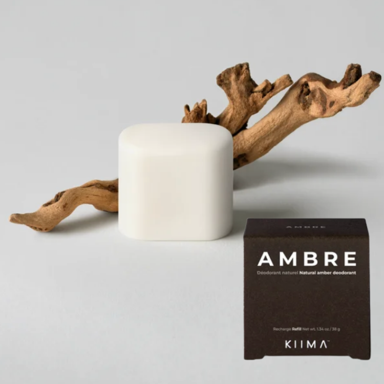 Bois d'ambre avec une recharge de déodorant arrondi et blanche et sa boite brune ou on peut lire Kiima Ambre déodorant naturel sur un fonc gris. 