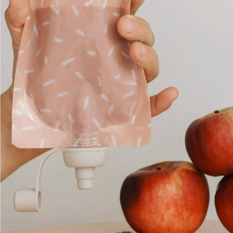 Pochette à boire réutilisable de la marque kangoo de couleur vieux-rose tenue par une main au-dessus de 3 pommes. 