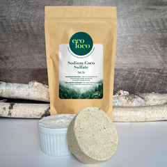 Pochette en papier kraft Eco Loco de Sodium Coco Sulfate SCS sur un fond de mur de bois avec deux branches de bouleaux et deux cake vaisselle dans un pot de céramique blanc