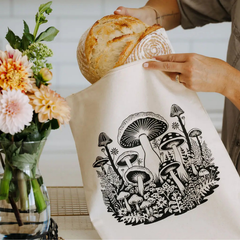 Une main qui met une miche de pain dans un sac à pain réutilisable de couleur naturelle avec des motifs de champignons.  À gauche du sac à pain, il y a un bouquet de fleur dans un pot en verre.