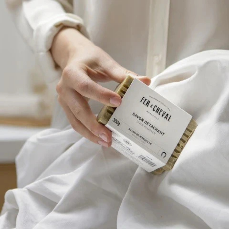Une barre de savon détachant verte avec une étiquette blanche écrit Fer à cheval 300 grammes tenu par une main de femme avec une chemise blanche au-dessus d'un tissu blanc. 