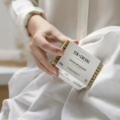 Une barre de savon détachant verte avec une étiquette blanche écrit Fer à cheval 300 grammes tenu par une main de femme avec une chemise blanche au-dessus d'un tissu blanc. 