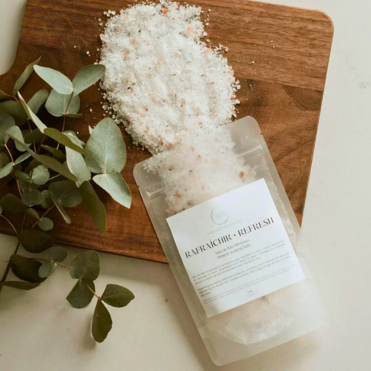 Un sac de sel de bain avec l'inscription rafraichir - refresh ouvert et se déversant sur une planche de bois.  Une plante se trouve sur la gauche et le fond est blanc. 